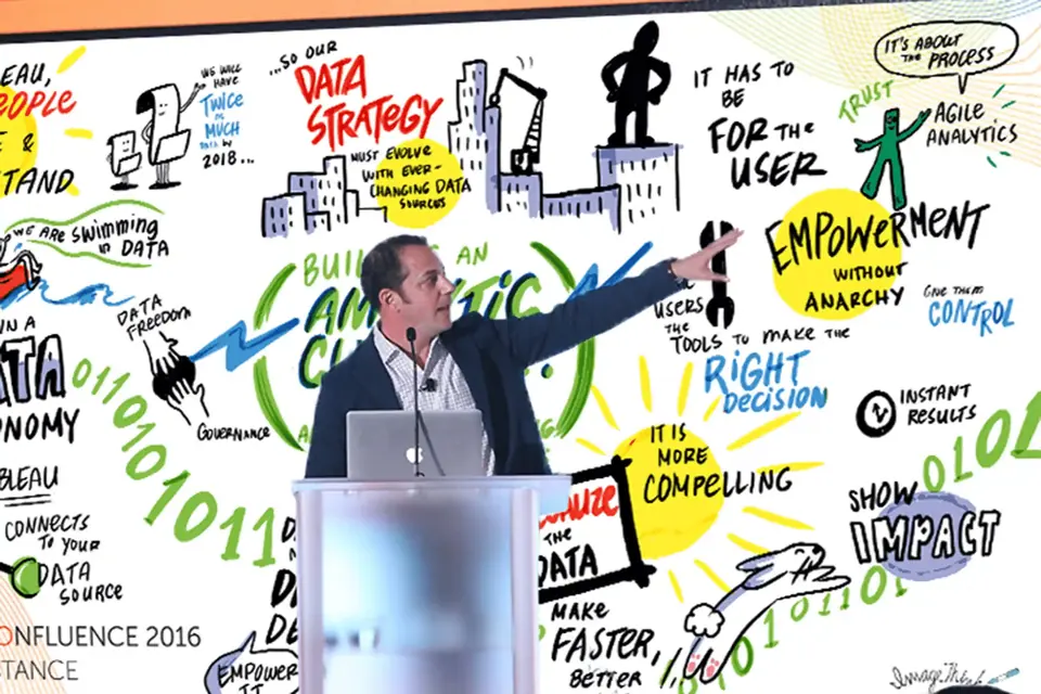 A man speaking at an event with a virtual scribing visual background.